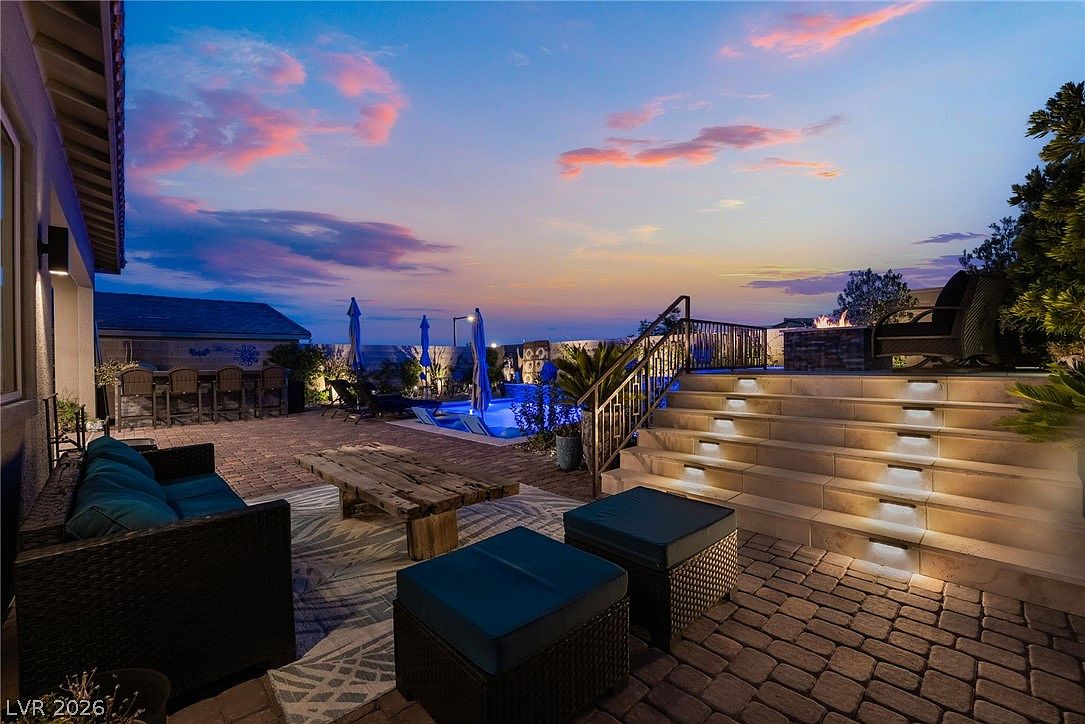 This inviting outdoor living space features a paved patio area furnished with a comfortable sofa and ottomans, perfect for evening relaxation. A set of illuminated stone steps leads up to an elevated deck area that includes a fire feature, while a swimming pool sits in the background under a vibrant sunset sky. The scene is framed by a modern metal railing and lush landscaping, creating a sophisticated and tranquil atmosphere for outdoor entertaining.