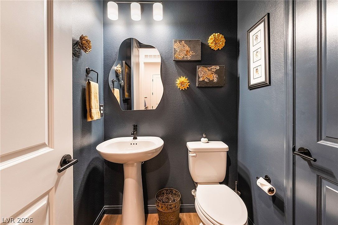 This compact powder room features bold, dark blue walls that create a dramatic and sophisticated atmosphere. The space is equipped with a white pedestal sink, a standard toilet, and an organic-shaped wall mirror, accented by decorative wall art and floral-inspired wall sculptures. A three-light vanity fixture provides illumination, while the wood-look flooring adds a touch of warmth to the moody color palette.