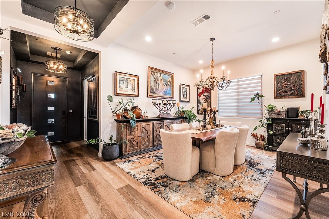 This elegant dining room features a large wooden table surrounded by six upholstered chairs, set atop a vibrant, abstract-patterned area rug. A rustic wooden sideboard provides ample storage, while a classic chandelier hangs above the table, complemented by recessed lighting and a decorative drum pendant in the adjacent entryway. The space is framed by warm-toned wood flooring and neutral walls, creating a sophisticated and inviting atmosphere.