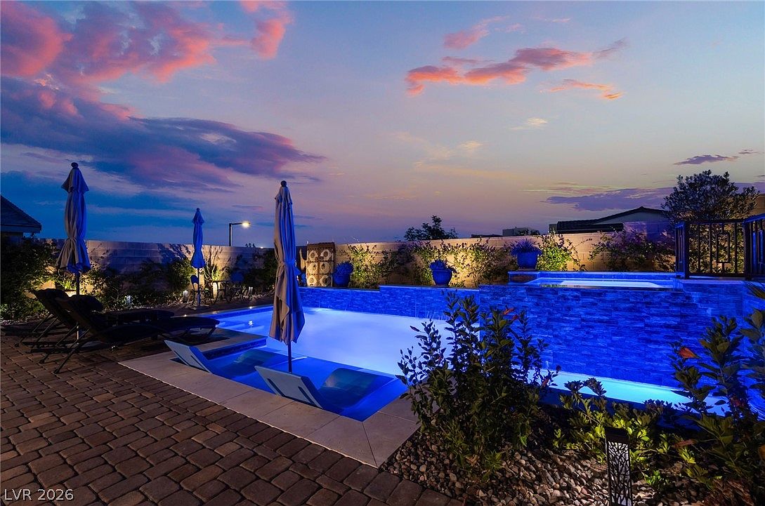 This stunning backyard oasis features a modern swimming pool with integrated lounge chairs and a raised spa, all illuminated by vibrant blue underwater lighting. The pool area is complemented by a paved patio, lounge chairs with umbrellas, and lush landscaping, set against a dramatic sunset sky. The perspective captures the serene and luxurious atmosphere of the outdoor living space at dusk.