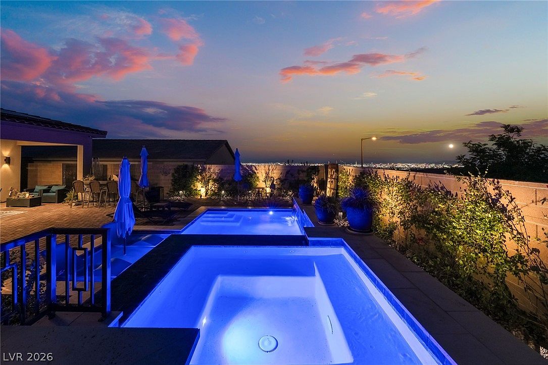 This stunning twilight view captures a modern backyard oasis featuring a tiered pool and spa illuminated by vibrant blue underwater lighting. The scene includes a paved patio area with outdoor dining furniture and umbrellas, set against a backdrop of a sunset sky and distant city lights. The composition emphasizes a luxurious, tranquil atmosphere perfect for evening relaxation and entertaining.