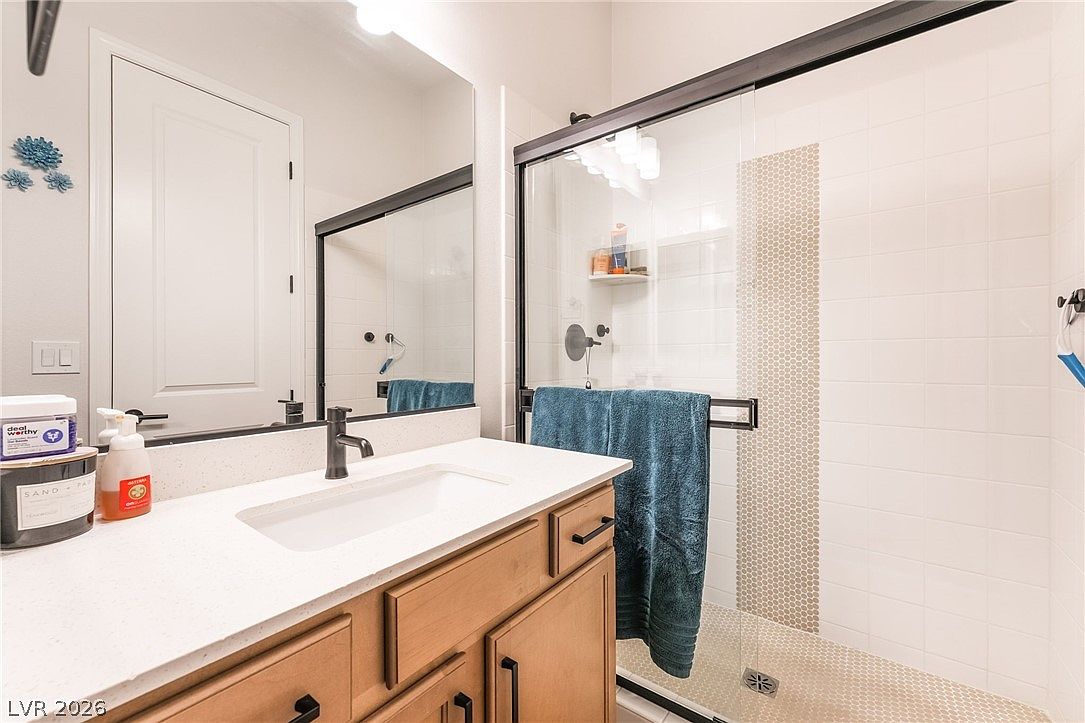 This bathroom features a light wood vanity with a white countertop and a single sink, paired with a large rectangular mirror. To the right, a glass-enclosed shower stall showcases a decorative vertical tile accent strip against white wall tiles and a textured floor. The space is bright and functional, offering a clean, modern aesthetic suitable for a guest or secondary bathroom.