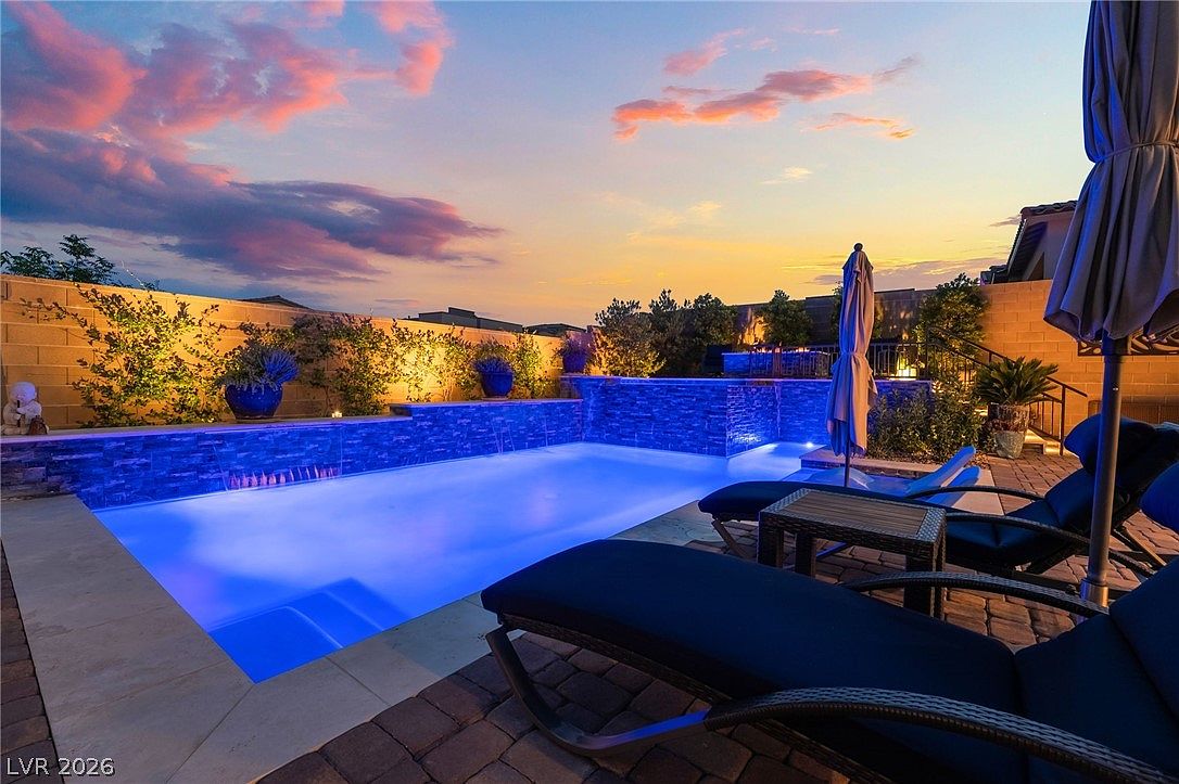 This backyard oasis features a rectangular swimming pool illuminated with vibrant blue underwater lighting, complemented by a stacked-stone water feature wall. The surrounding patio is paved with dark stone pavers and includes lounge chairs with umbrellas, creating a relaxing atmosphere. The scene is captured at sunset, with a dramatic, colorful sky providing a picturesque backdrop to the well-manicured outdoor living space.