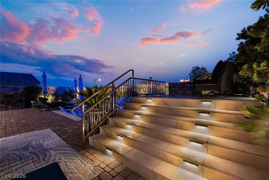 This outdoor living space features a set of stone steps illuminated by integrated stair lighting, leading up to a raised patio area. The scene is captured at dusk, showcasing a vibrant sunset sky above a pool area with lounge chairs and umbrellas. The combination of warm lighting and the twilight atmosphere creates an inviting and luxurious ambiance for evening relaxation.