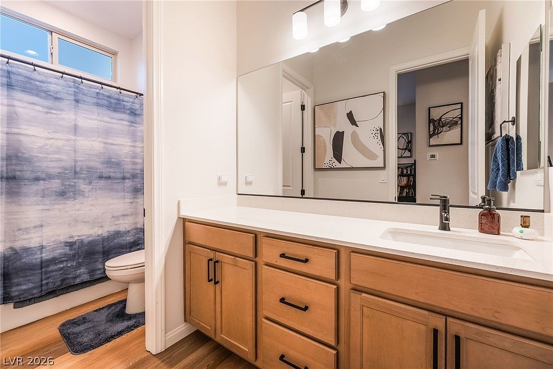 This bathroom features a light wood vanity with dark hardware, topped with a white countertop and a single sink. A large, frameless mirror spans the wall above the vanity, reflecting the adjacent bedroom area and a piece of abstract wall art. To the left, a blue and white patterned shower curtain conceals the tub and toilet area, while the room is finished with wood-look flooring.