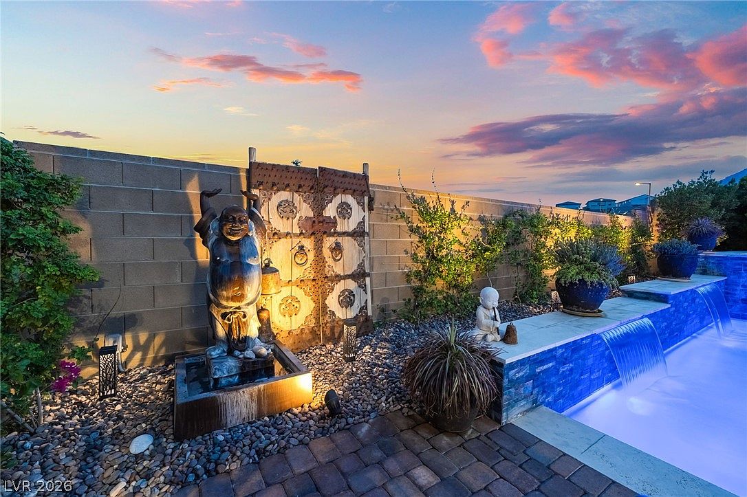 This serene backyard oasis features a modern swimming pool with a cascading water feature and a decorative stone wall adorned with a large, weathered wooden gate and a prominent Buddha statue. The area is landscaped with river rock, potted plants, and a small meditative statue, all set against a vibrant sunset sky. The perspective is a ground-level shot that captures the tranquil, resort-like atmosphere of the outdoor living space.