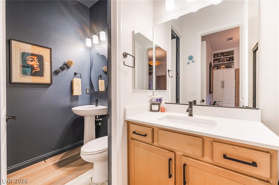 This image captures a dual-space bathroom layout, featuring a powder room with a dark blue accent wall and a pedestal sink, adjacent to a vanity area with light wood cabinetry and a white countertop. A large mirror reflects the vanity and a glimpse into an adjoining room, while modern light fixtures provide bright illumination. The combination of the bold wall color and the neutral vanity creates a contemporary and functional aesthetic.