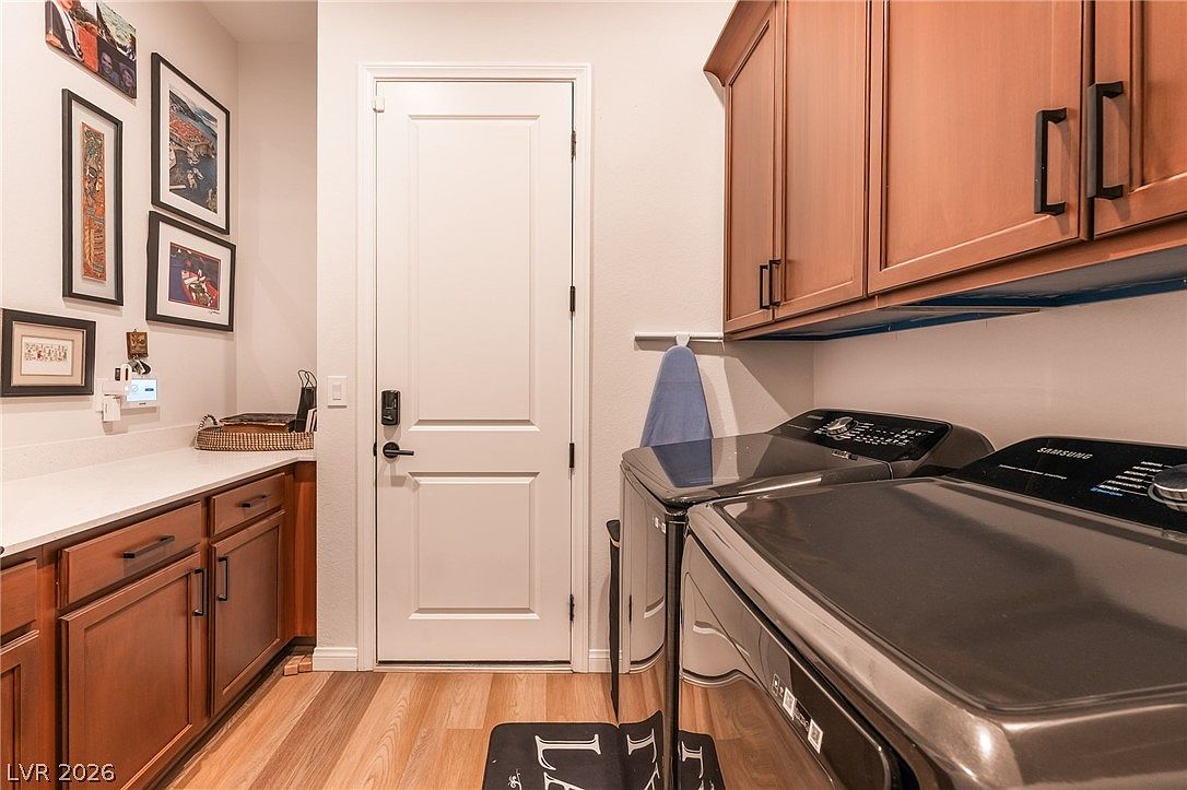 This functional laundry room features dark-toned, high-gloss appliances paired with warm-toned wood cabinetry and light-colored countertops. The space includes a central door, a wall-mounted ironing board, and a gallery wall of framed artwork, creating a tidy and organized atmosphere. The perspective is a straight-on view that highlights the efficient use of space and the contrast between the dark appliances and the neutral wall color.