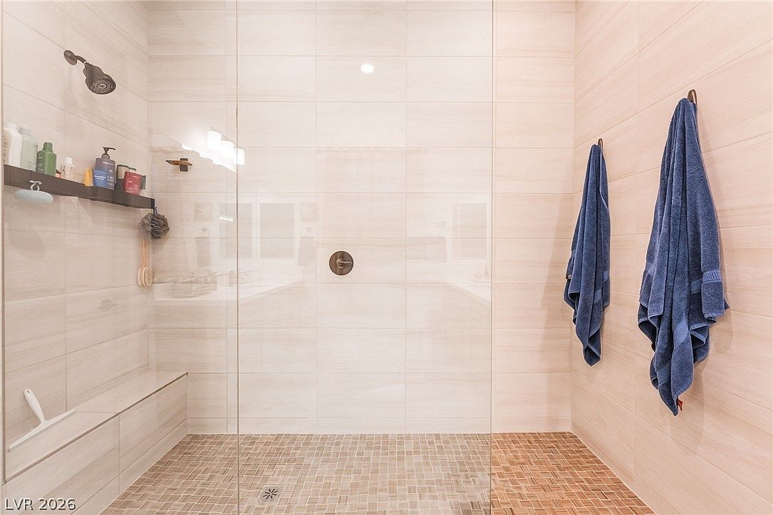 This modern walk-in shower features large, light-toned rectangular wall tiles and a textured, small-format tile floor. A glass partition separates the shower area, which includes a built-in bench, a wall-mounted shelf for toiletries, and a shower head. Two blue towels hang on the right wall, adding a pop of color to the neutral, spa-like space.