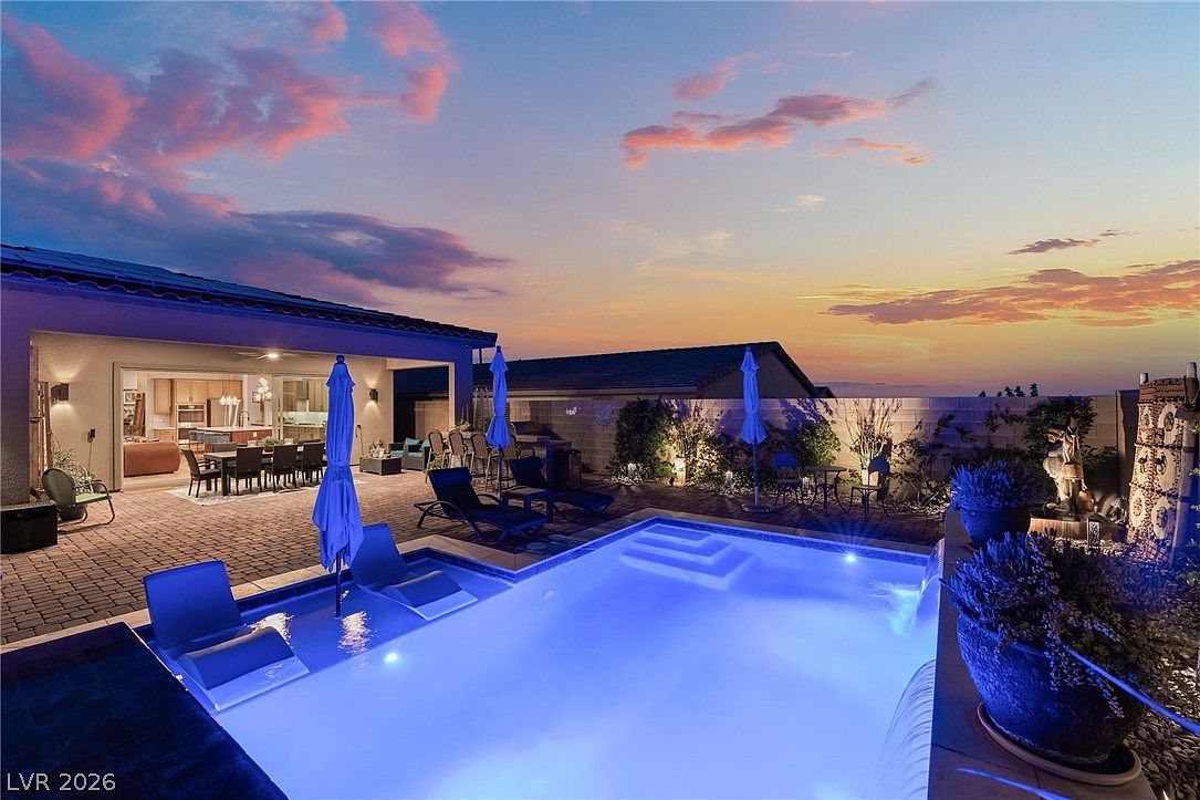 This stunning backyard oasis features a modern swimming pool with a built-in tanning ledge and a cascading water feature, set against a vibrant sunset sky. The paved patio area includes comfortable lounge chairs and a dining set, seamlessly connecting to the home's open-concept interior through large sliding glass doors. The space is thoughtfully landscaped with potted plants and ambient lighting, creating a luxurious and inviting atmosphere for evening relaxation.