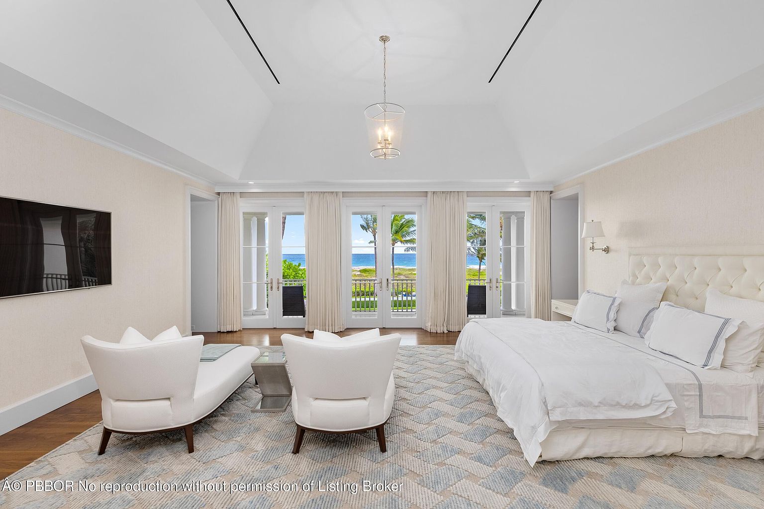 This is a bright and airy primary bedroom featuring a king-size bed with white linens and a tufted headboard. The room has a seating area with two white chairs and a modern glass side table. French doors lead to a balcony with ocean views, enhancing the luxurious and serene atmosphere.