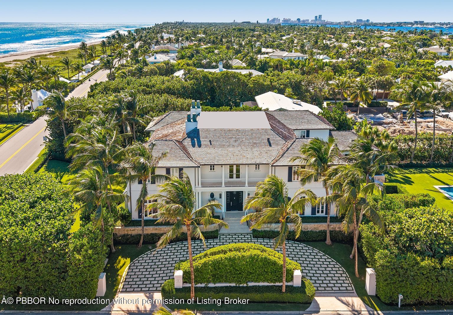 This aerial view showcases a grand, two-story white house with a symmetrical facade, a dark tile roof, and multiple chimneys. The property is surrounded by lush greenery, including mature palm trees and manicured hedges, with a patterned driveway leading to the entrance. In the background, the ocean and a distant cityscape add to the property's desirable location and luxurious appeal.
