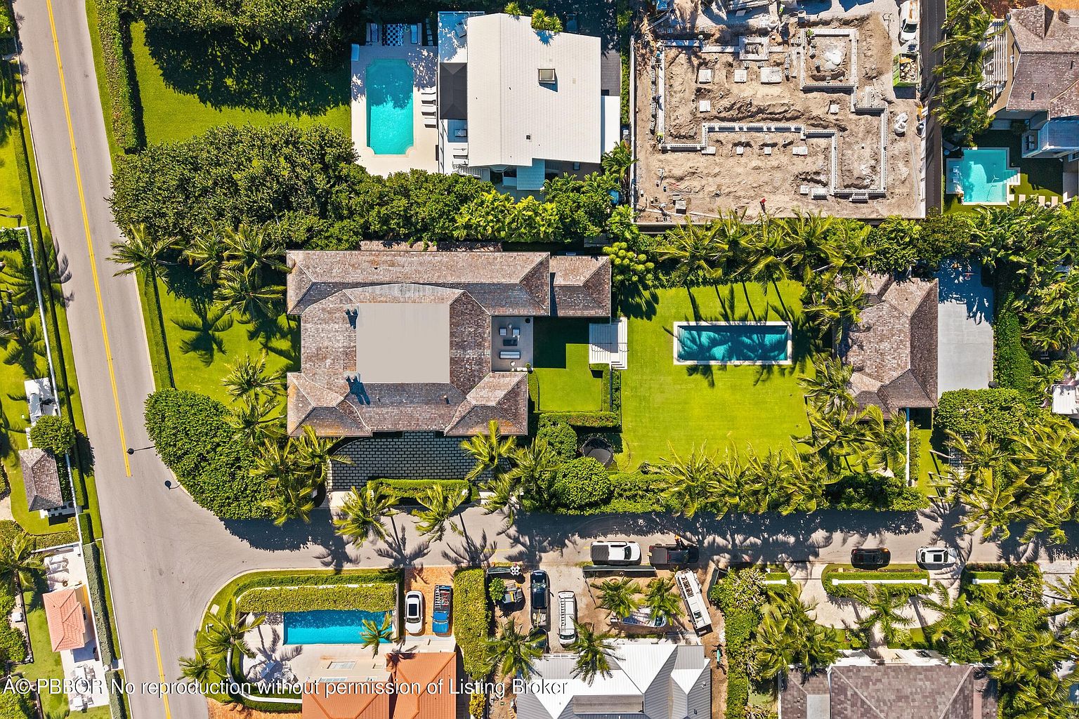 This aerial view showcases a luxurious residential property featuring multiple structures, including a main house with a distinctive roof, a separate pool house, and a large swimming pool. Lush landscaping, including palm trees and manicured lawns, surrounds the buildings, creating a private and serene environment. A construction site is visible nearby, indicating ongoing development in the area.