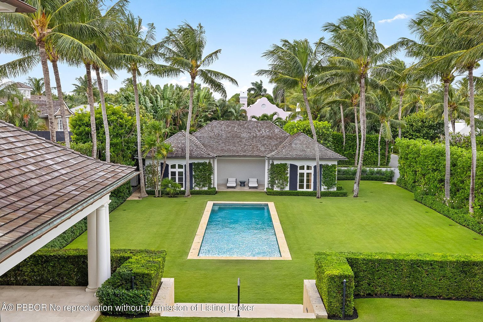 This is an aerial view of a luxurious backyard featuring a rectangular swimming pool surrounded by lush green grass and manicured hedges. A pool house with a dark shingle roof and white trim sits at the far end of the pool, flanked by tall palm trees. The scene evokes a sense of tranquility and upscale living.