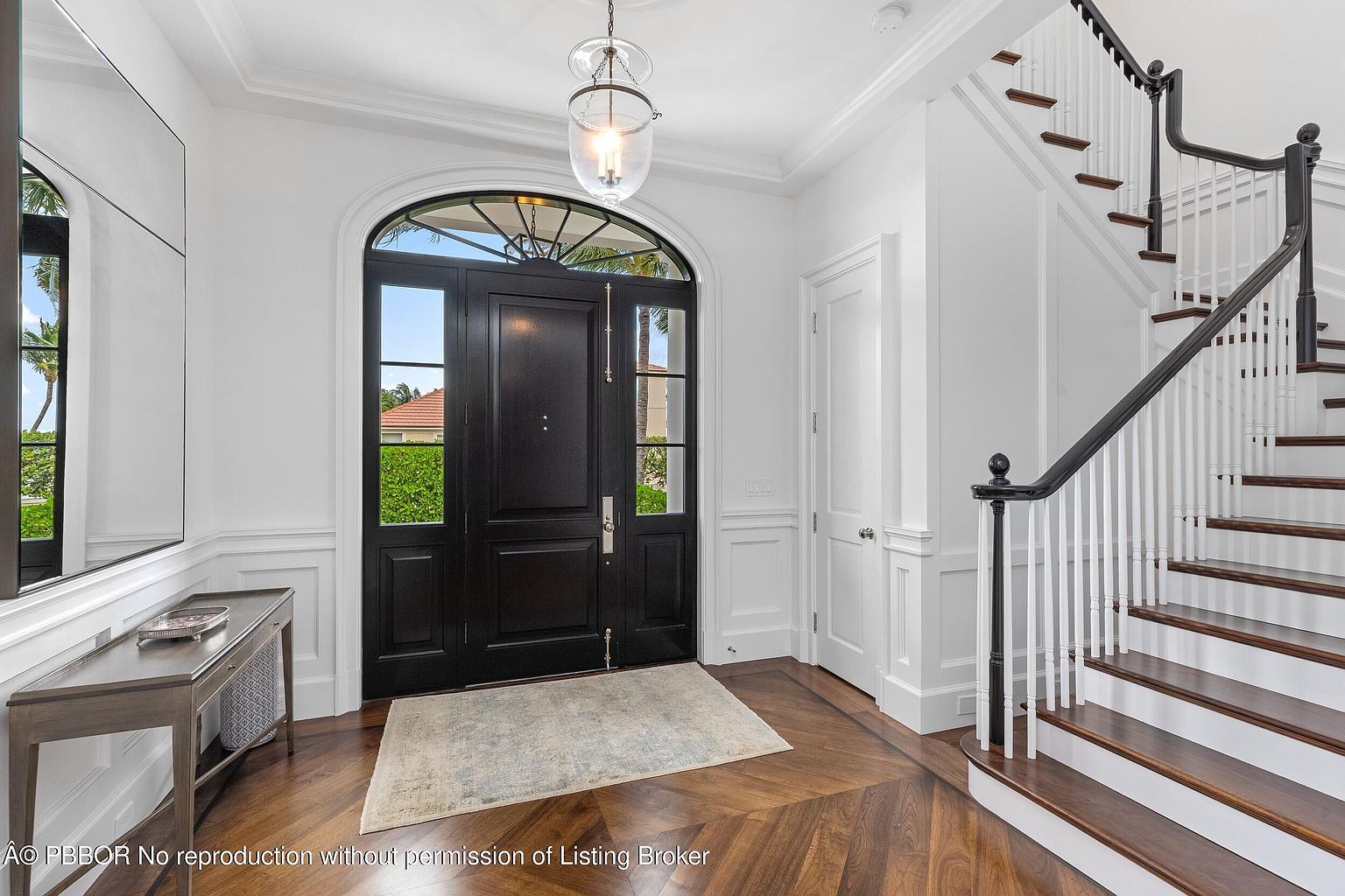 This is an interior shot of a grand foyer featuring a dark wood herringbone floor, a black front door with glass panels, and a staircase with dark wood treads and a black handrail. The walls are painted white with decorative molding, and a console table sits to the left of the door. The overall impression is elegant and luxurious.