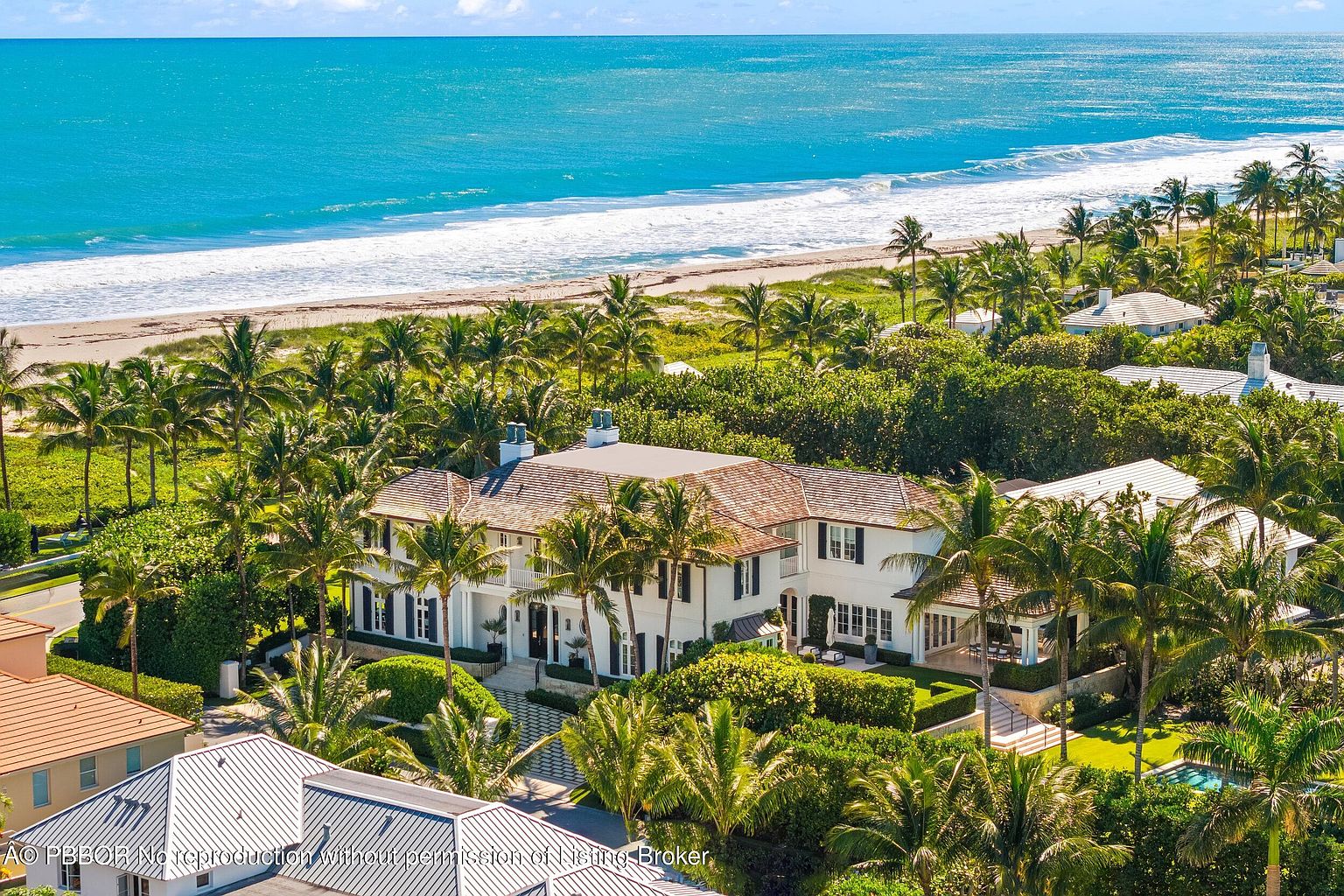 This aerial view showcases a luxurious beachfront property with a grand two-story house surrounded by lush tropical landscaping, including numerous palm trees and manicured hedges. The house features a light-colored exterior, a tile roof, and multiple windows, offering stunning ocean views. The turquoise ocean and sandy beach create a picturesque backdrop, emphasizing the property's prime location and upscale appeal.