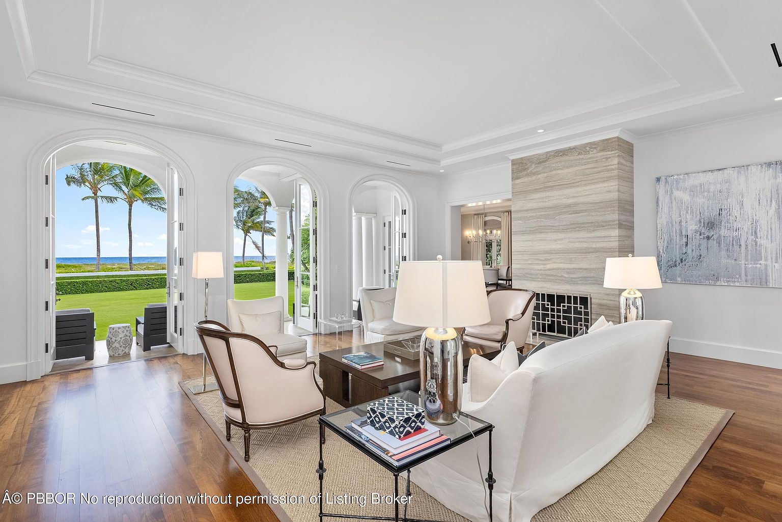 This is an interior shot of a luxurious living room featuring a neutral color palette with white walls and light-colored furniture. The room is well-lit with natural light streaming in through large arched doorways that offer views of the ocean and palm trees. A fireplace with a stone surround adds a focal point, and the hardwood floors and area rug contribute to the room's elegant and comfortable atmosphere.
