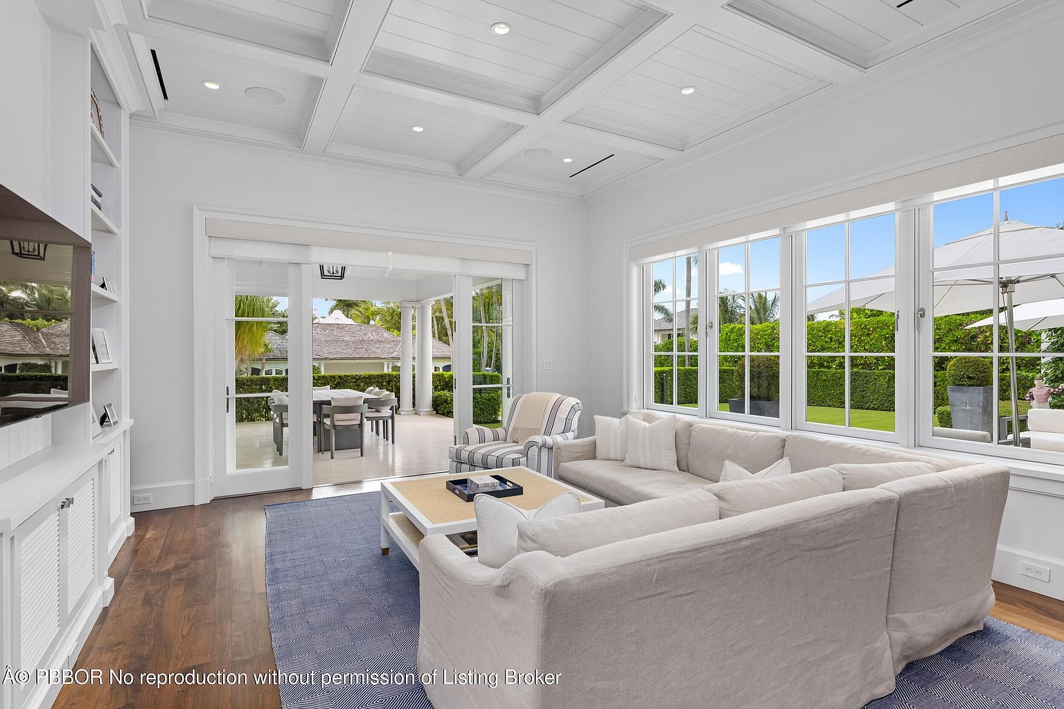 This is a bright and airy living room featuring a large, comfortable sectional sofa, a stylish coffee table, and a blue rug. The room is bathed in natural light from the expansive windows overlooking the outdoor space, which includes a patio and lush greenery. The coffered ceiling and white walls contribute to the room's elegant and inviting atmosphere.