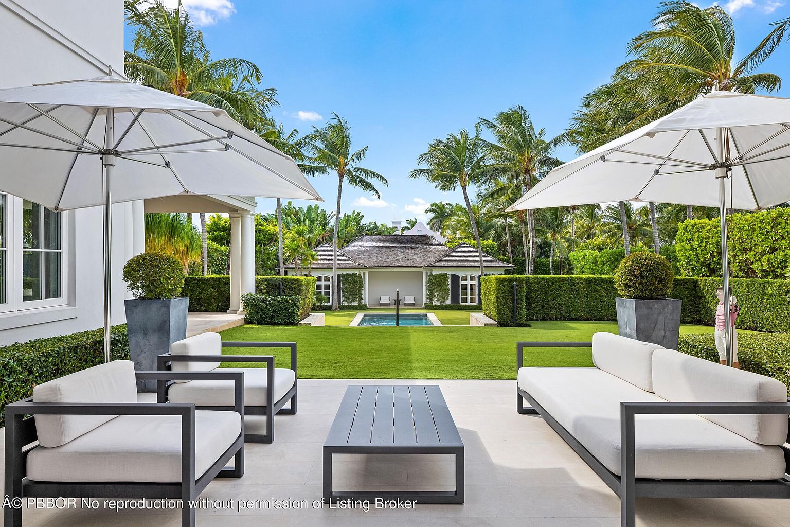 This image showcases a beautifully landscaped backyard featuring a well-manicured lawn, trimmed hedges, and mature palm trees. A pool house with a dark roof and white trim is centered in the background, complemented by a rectangular pool. Modern outdoor furniture, including sofas, chairs, and a coffee table, is arranged on a tiled patio, creating an inviting outdoor living space.