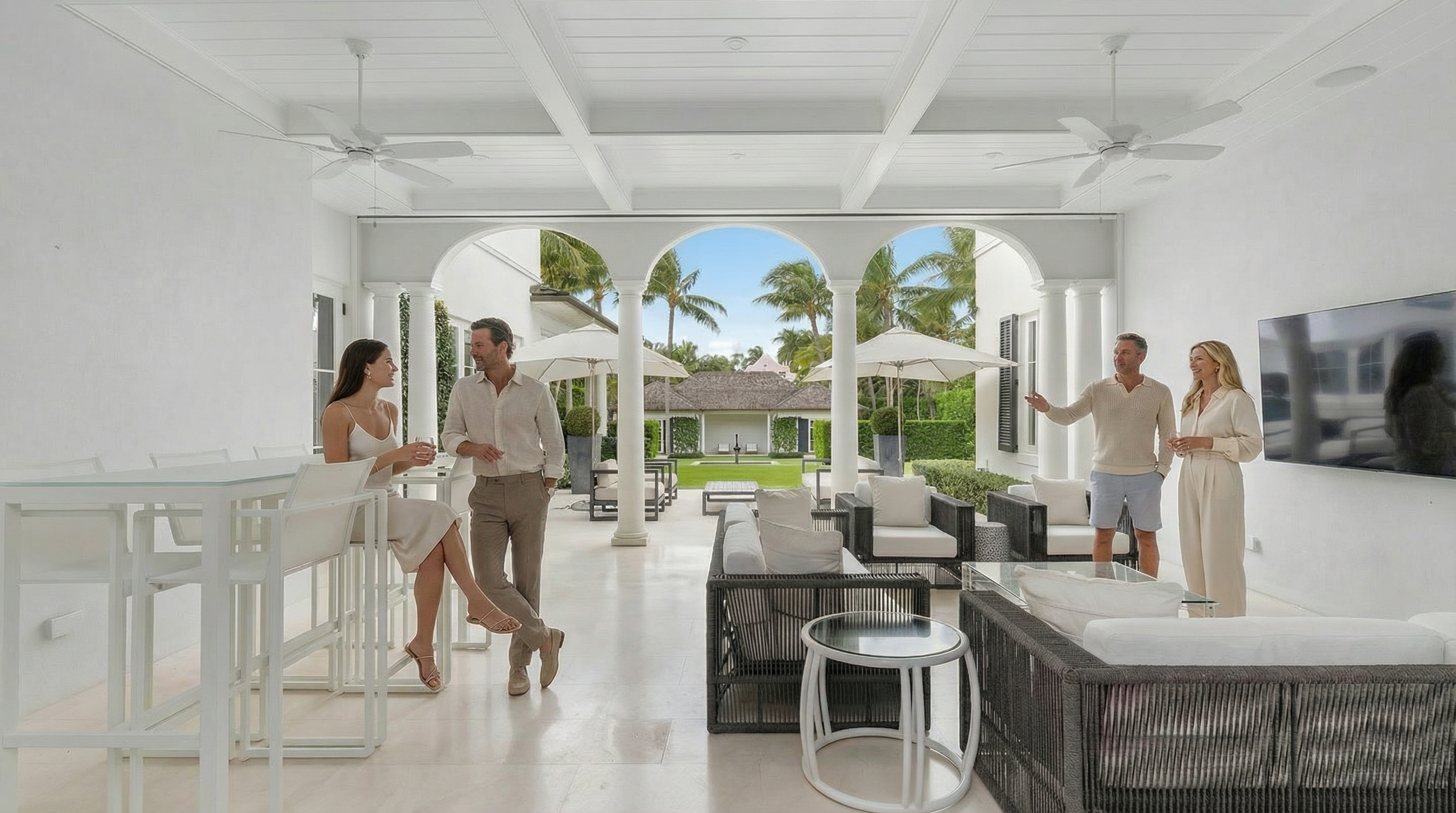 This luxurious covered outdoor patio space features sophisticated modern furniture and an open-arch design that flows seamlessly into the tropical backyard. The area is equipped with ceiling fans and a wall-mounted flat-screen television, perfect for relaxation and social gatherings. Its bright, white-toned palette and spacious layout create an elegant, airy atmosphere suitable for upscale outdoor living.