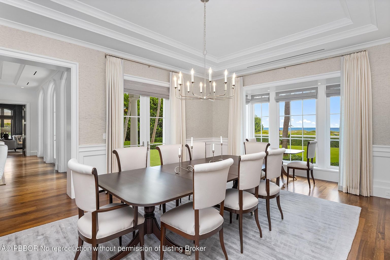 This is an interior shot of a dining room featuring a dark wood dining table surrounded by eight upholstered chairs with wooden frames. A modern chandelier hangs above the table, and natural light floods the room through large windows dressed with sheer curtains. The room is decorated in neutral tones with hardwood floors and a light-colored area rug, creating an elegant and inviting atmosphere.
