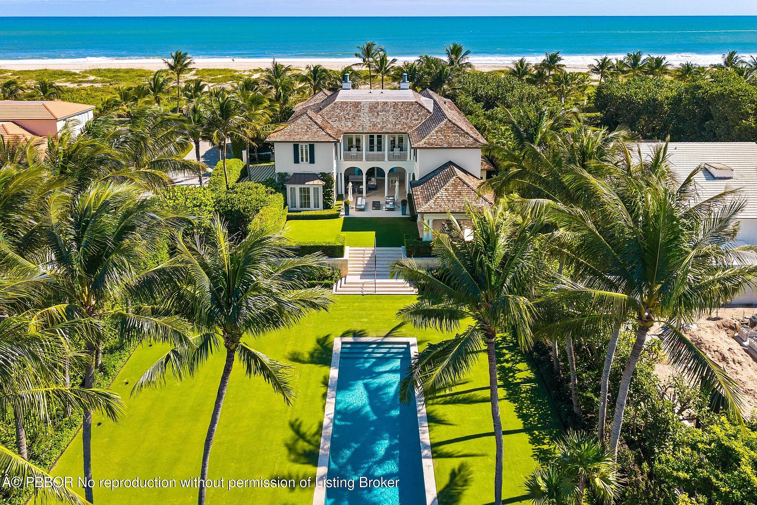 This aerial shot showcases a luxurious beachfront property with a grand house, a long rectangular swimming pool, and meticulously manicured lawns framed by lush palm trees. The house features a light-colored facade, a multi-level design, and a tiled roof, exuding an air of elegance and exclusivity. The ocean is visible in the background, emphasizing the prime location.