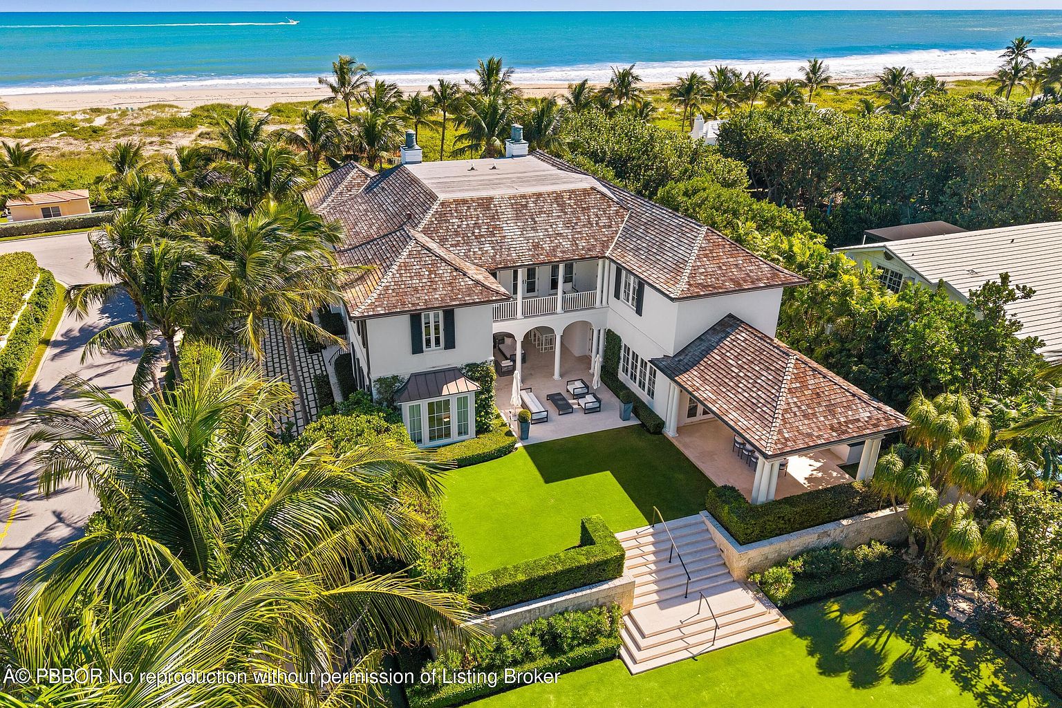 This aerial view showcases a luxurious beachfront property with a meticulously maintained lawn and lush landscaping. The house features a distinctive brown tile roof, white exterior walls, and a charming patio area with outdoor seating. The property offers direct access to a pristine beach and the turquoise ocean, creating an idyllic coastal living experience.