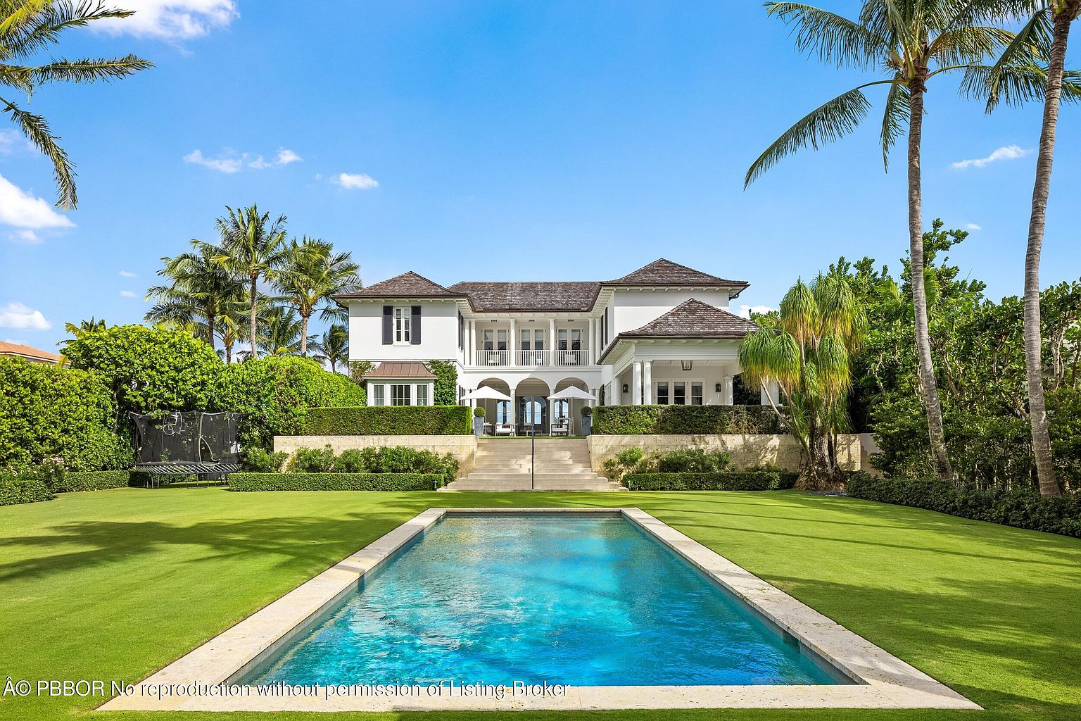 This image showcases a luxurious backyard featuring a rectangular pool with a light stone border, surrounded by a lush green lawn. The pool reflects the clear blue sky, enhancing the serene atmosphere. In the background, a grand white house with dark shutters and a brown roof is partially visible, framed by palm trees and manicured hedges, suggesting a high-end property.