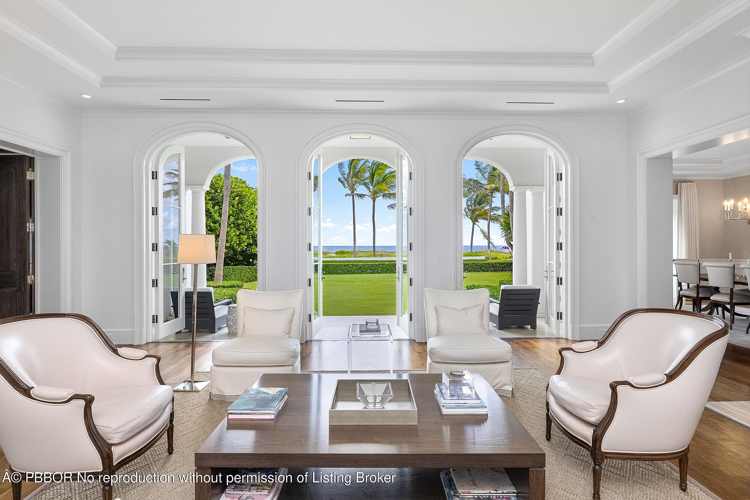 This is an interior shot of a luxurious living room featuring three arched doorways that open to a lush green lawn and ocean view. The room is furnished with elegant white armchairs and a large dark wood coffee table, creating a sophisticated and inviting atmosphere. The overall style is classic and refined, emphasizing the connection between indoor and outdoor spaces.