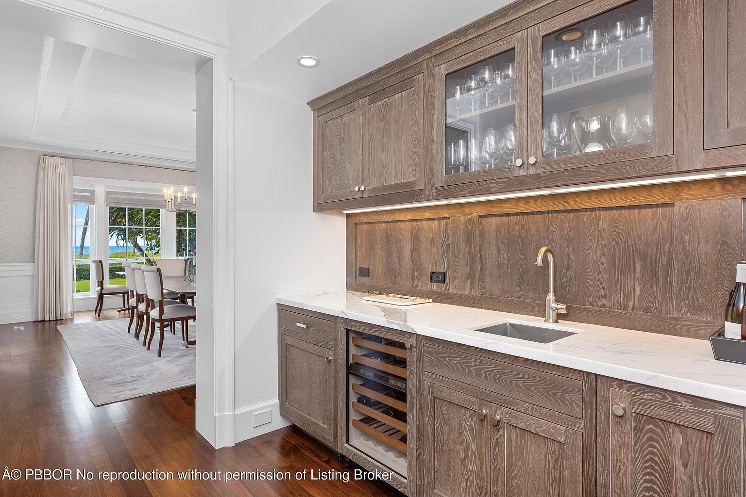 This interior shot showcases a sophisticated kitchen area with custom wood cabinetry, glass-fronted upper cabinets displaying glassware, and a marble countertop. A built-in wine cooler adds a touch of luxury, while a stainless steel sink and faucet provide functionality. The open doorway leads to a dining area with a view of the ocean, enhancing the home's appeal.