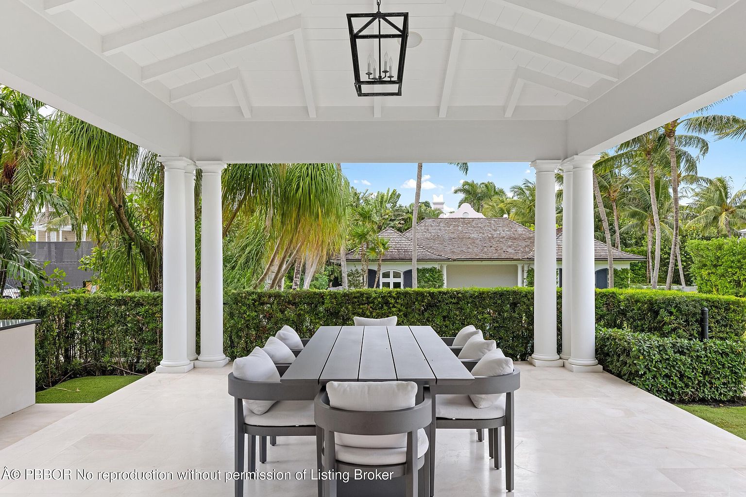 This image showcases a beautifully designed outdoor patio area, featuring a large dining table with comfortable seating, perfect for entertaining. The space is covered by a white, well-maintained roof supported by elegant columns, providing shade and a sense of grandeur. Lush greenery surrounds the patio, creating a serene and private atmosphere, while a glimpse of a charming house in the background adds to the overall appeal.