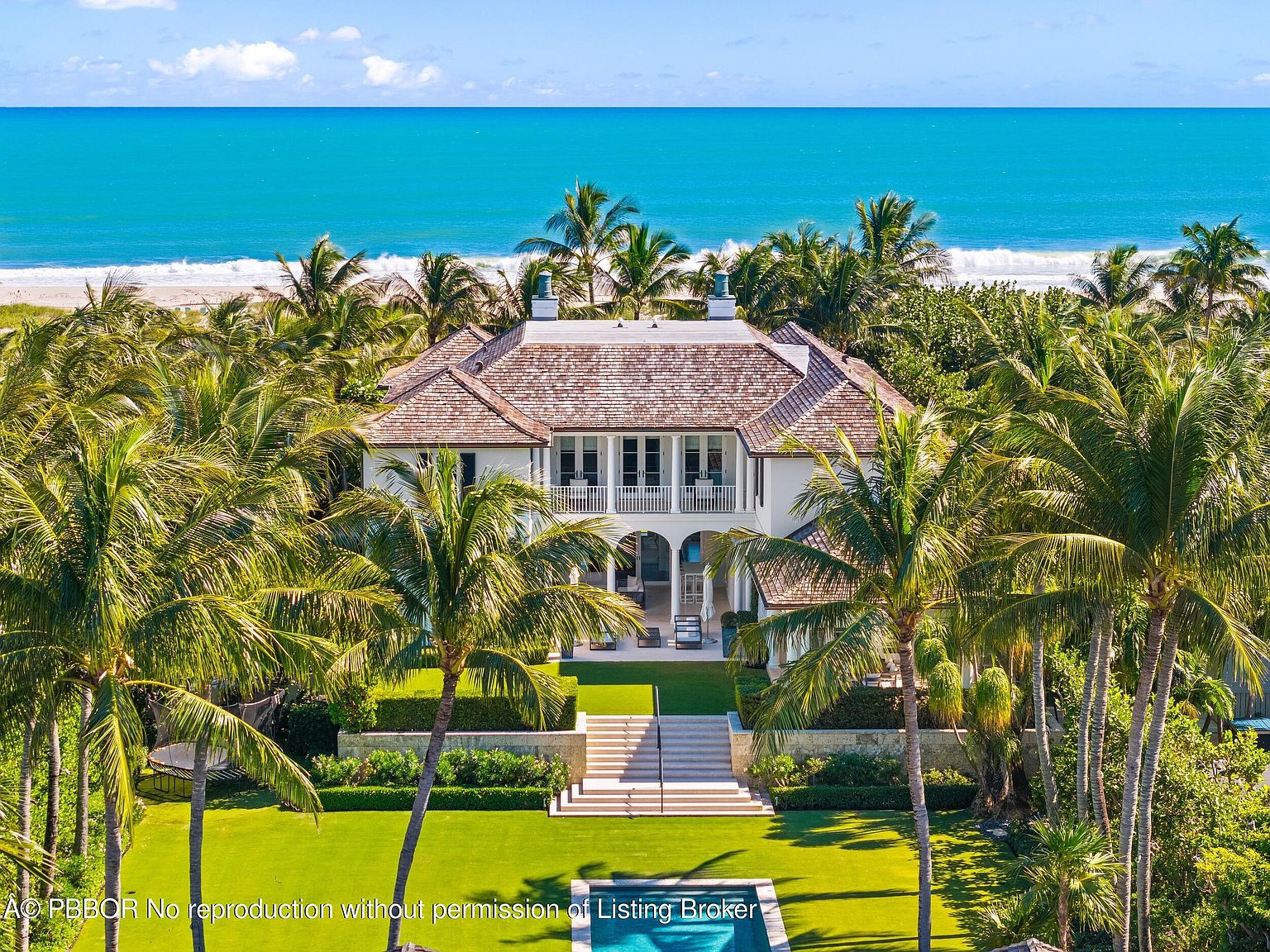 This aerial view showcases a luxurious beachfront property surrounded by lush palm trees. The house features a light-colored exterior, a multi-level design with balconies, and a well-manicured lawn leading to a swimming pool. The turquoise ocean provides a stunning backdrop, emphasizing the property's prime location and upscale appeal.