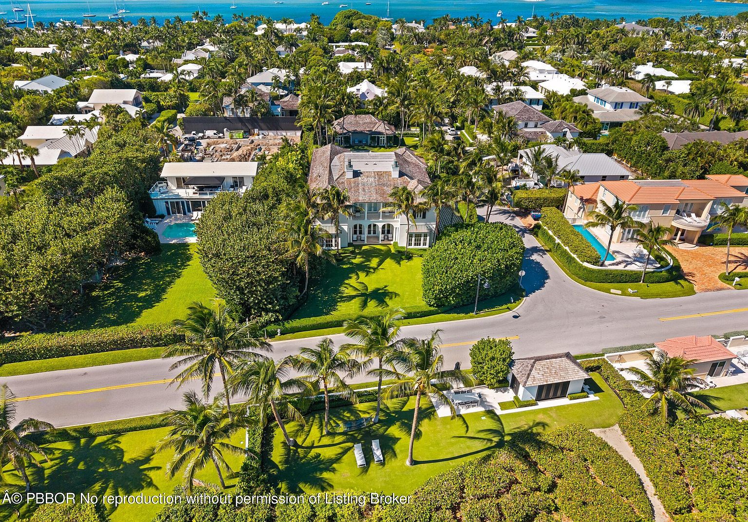 This aerial shot showcases a luxurious residential area with lush greenery and several upscale homes. Palm trees line the streets and surround the properties, adding a tropical feel. The houses feature varying architectural styles, some with pools and well-manicured lawns, all set against a backdrop of a beautiful blue ocean.