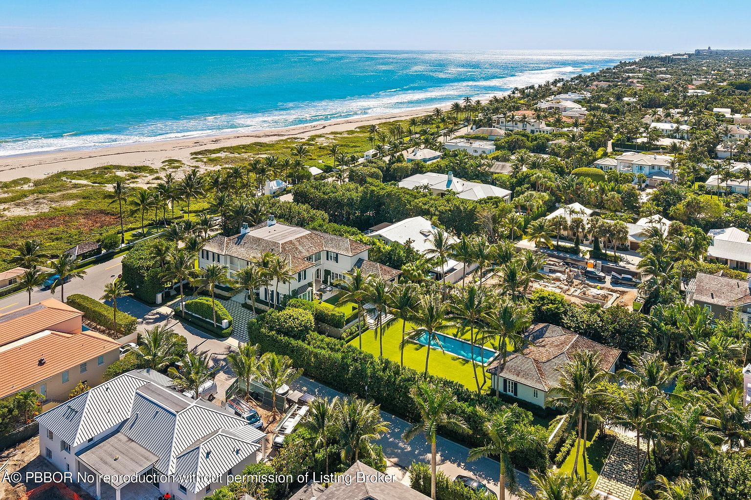 This aerial shot showcases a luxurious coastal property with lush landscaping, including palm trees and manicured hedges. A sparkling blue swimming pool is a focal point, complemented by the nearby beach and ocean views. The overall impression is one of upscale coastal living and privacy.