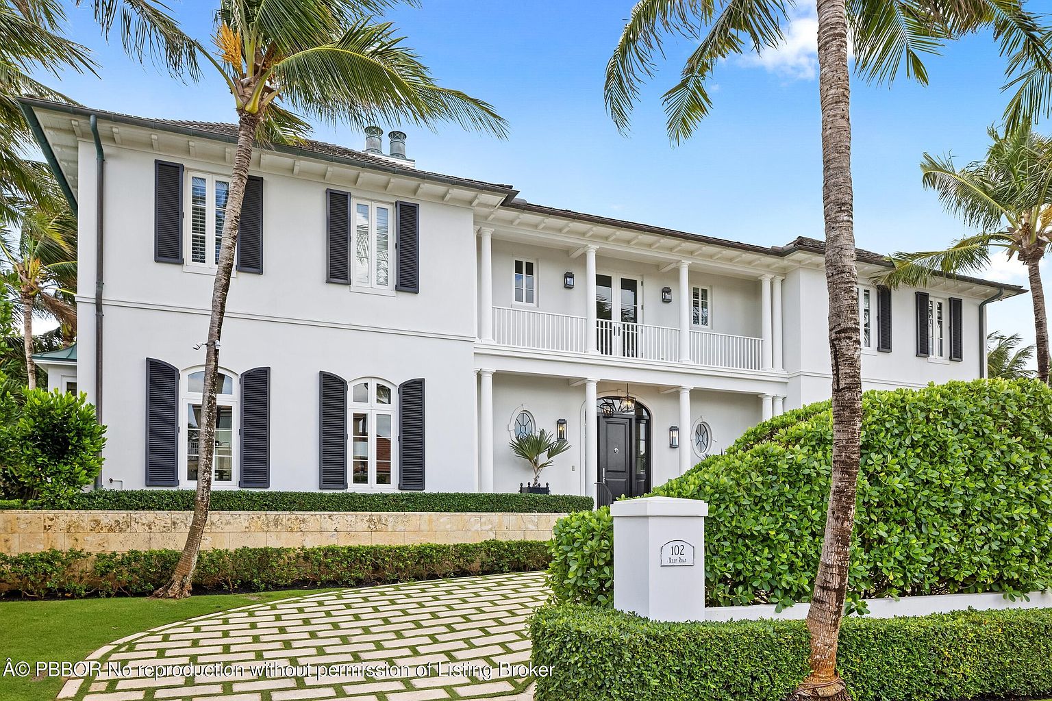 This is a front exterior view of a large, elegant two-story house with a white facade and dark shutters. The property features a well-manicured lawn, a patterned driveway, and lush green hedges. A covered balcony with white columns adds to the home's stately appearance, and palm trees frame the scene, suggesting a tropical or coastal location.