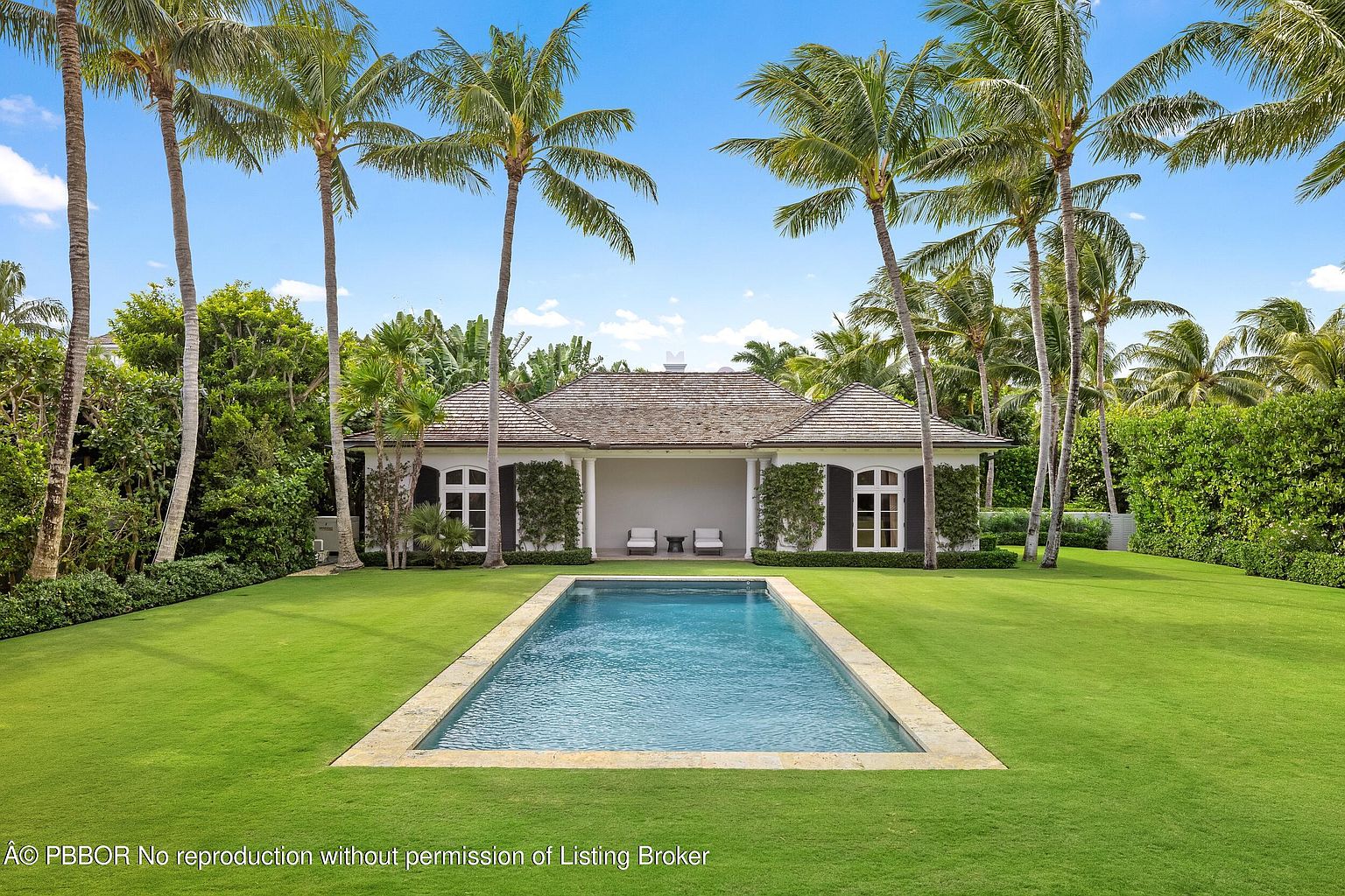 This image showcases a luxurious backyard featuring a rectangular swimming pool with a stone border, surrounded by a lush green lawn. A charming pool house with dark shutters and a light-colored roof stands at the far end of the pool, framed by tall palm trees and manicured hedges. The scene evokes a sense of tranquility and upscale outdoor living.