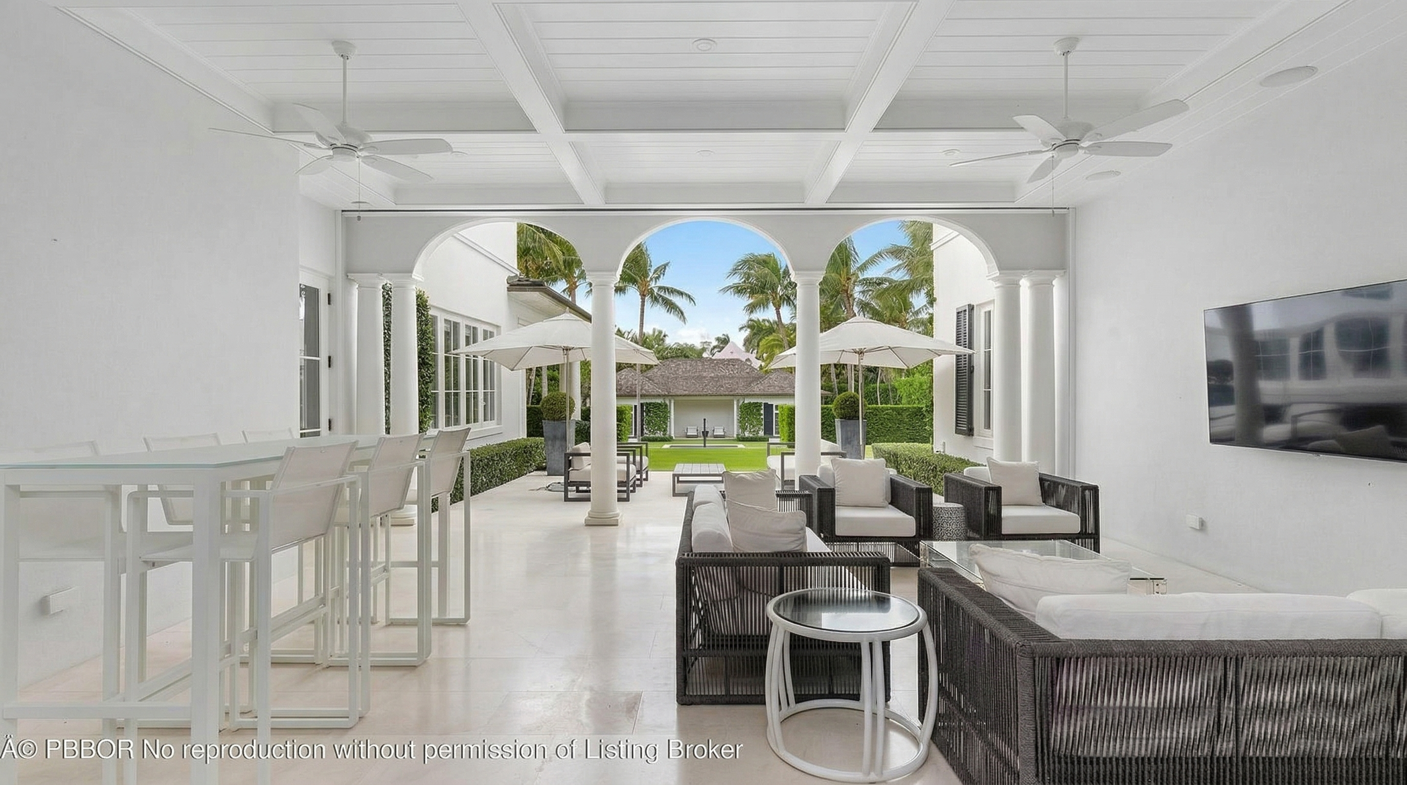 This image showcases a luxurious outdoor living space, featuring a covered patio with elegant white columns and ceiling beams. The patio is furnished with comfortable seating arrangements, including woven sofas and armchairs, complemented by sleek side tables. Through the arched openings, a beautifully landscaped backyard with palm trees and additional seating areas is visible, creating a seamless transition between indoor and outdoor living.