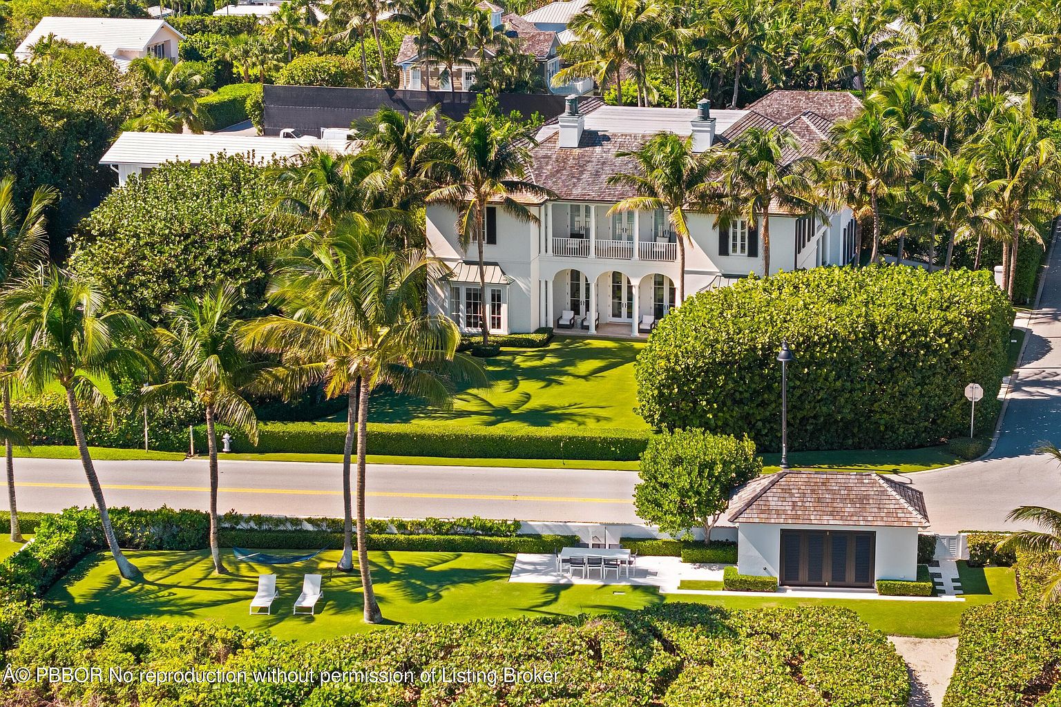 This aerial view showcases a luxurious estate with meticulously manicured lawns, lush tropical landscaping, and a grand white residence featuring a balcony and arched entryways. A separate structure with a wooden roof adds to the property's charm, while palm trees line the street, enhancing the overall curb appeal and creating a serene, upscale ambiance.