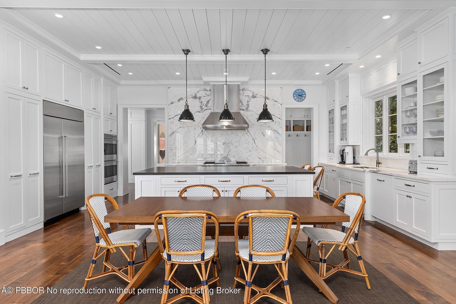This is a bright and spacious kitchen featuring white cabinetry, stainless steel appliances, and a large island with a dark countertop. The kitchen also includes a dining area with a wooden table and woven chairs. The overall impression is one of modern elegance and functionality, perfect for both cooking and entertaining.