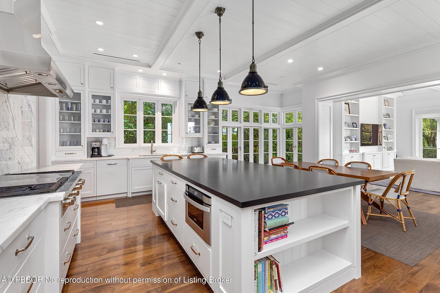 This is a bright and spacious kitchen featuring white cabinetry, a dark countertop island with built-in shelving, and stainless steel appliances. Natural light floods the space through large windows, highlighting the hardwood floors and open layout that connects to the dining area. The overall impression is one of modern elegance and functionality.