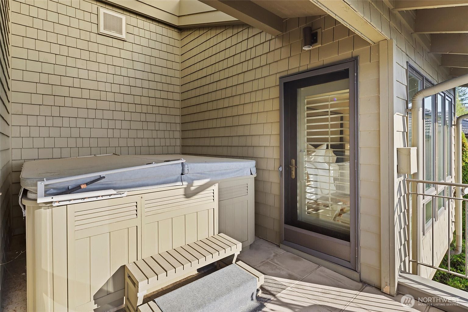 This image showcases a private outdoor patio area featuring a covered hot tub with a matching step stool, set against light-colored shingle siding. A glass-paned door provides direct access to the interior, while a metal railing borders the edge of the space. The scene offers a cozy, relaxing atmosphere perfect for a residential listing.