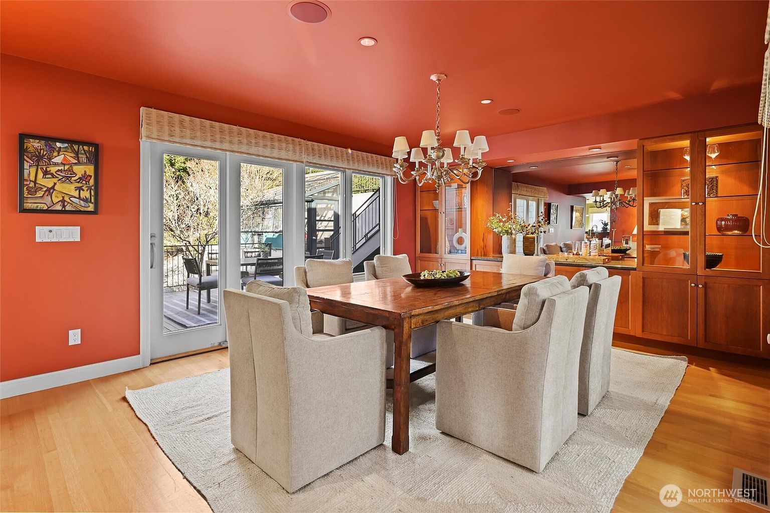 This dining room features a warm, inviting atmosphere with vibrant orange walls and a rich wooden dining table surrounded by six upholstered chairs. A classic chandelier hangs above the table, while a large built-in wooden hutch provides elegant storage and display space. The room is brightened by a wall of glass doors that open onto an outdoor deck, creating a seamless transition between indoor and outdoor living.