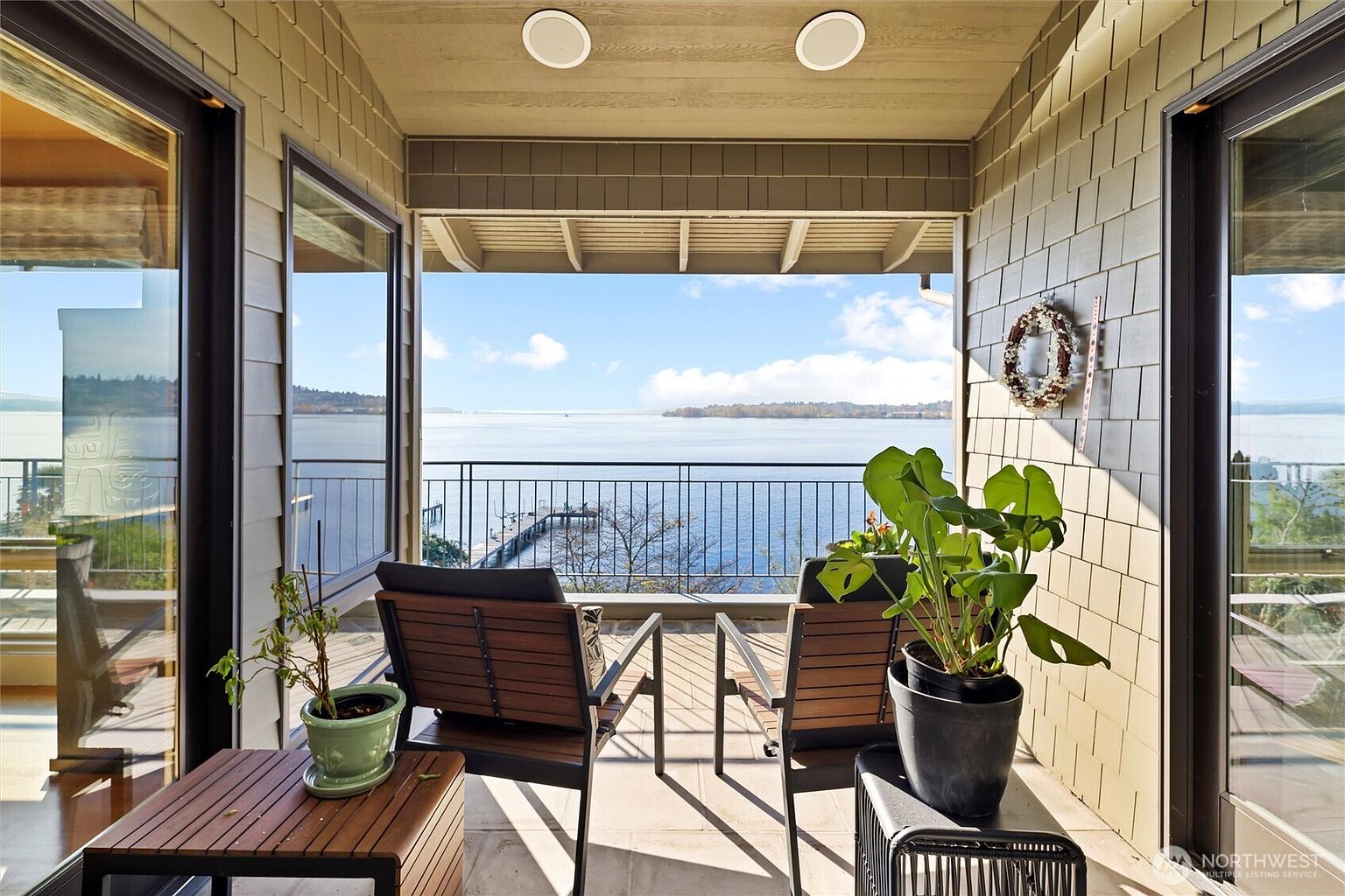 This covered balcony offers a serene, elevated view of a calm lake and a private dock, framed by shingled exterior walls and large glass doors. The space is furnished with two wooden chairs and potted plants, creating a peaceful retreat for relaxation. The perspective is from inside the covered area looking out, emphasizing the seamless transition between indoor comfort and the expansive waterfront scenery.