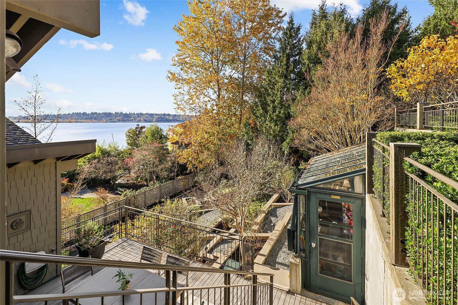 This elevated view captures a multi-level outdoor living space featuring a wooden deck with a dining area and a glass-enclosed sunroom or greenhouse structure. The scene overlooks a lush, terraced garden that leads down toward a serene lake, framed by mature trees with autumn foliage. The perspective provides a cinematic look at the home's integration with its natural, waterfront surroundings.