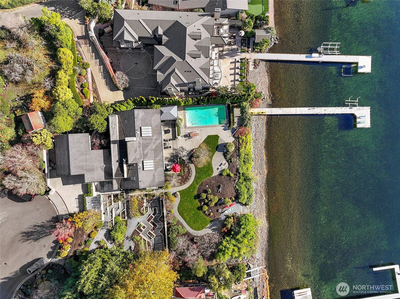 This high-angle aerial view captures a luxurious waterfront estate featuring two distinct residential structures, a private swimming pool, and two long docks extending into the clear lake. The property is surrounded by lush, mature landscaping, winding pathways, and a well-manicured lawn that transitions into a rocky shoreline. The perspective emphasizes the expansive scale of the property and its prime location directly on the water's edge.