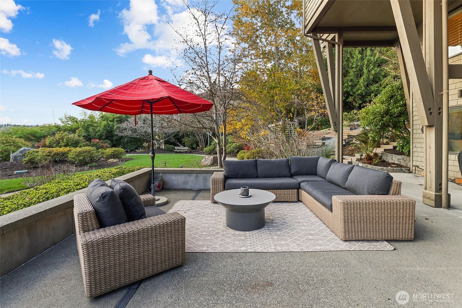 This spacious concrete patio features a comfortable, modern wicker sectional sofa with dark grey cushions and a matching armchair, centered around a sleek, circular coffee table. A vibrant red umbrella provides shade, while a patterned area rug defines the seating space, all set against a backdrop of a lush, landscaped backyard and a clear blue sky. The perspective is a wide, eye-level shot that emphasizes the inviting outdoor living area and its seamless connection to the surrounding greenery.