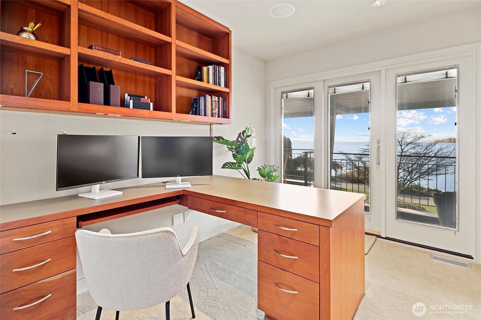 This bright and professional home office features a large, L-shaped wooden desk with built-in cabinetry and shelving, providing ample workspace and storage. The room is anchored by a modern, light-colored upholstered chair and dual computer monitors, while large glass doors offer a stunning, unobstructed view of the water and outdoor scenery. The clean lines and warm wood tones create a productive yet serene atmosphere perfect for remote work.