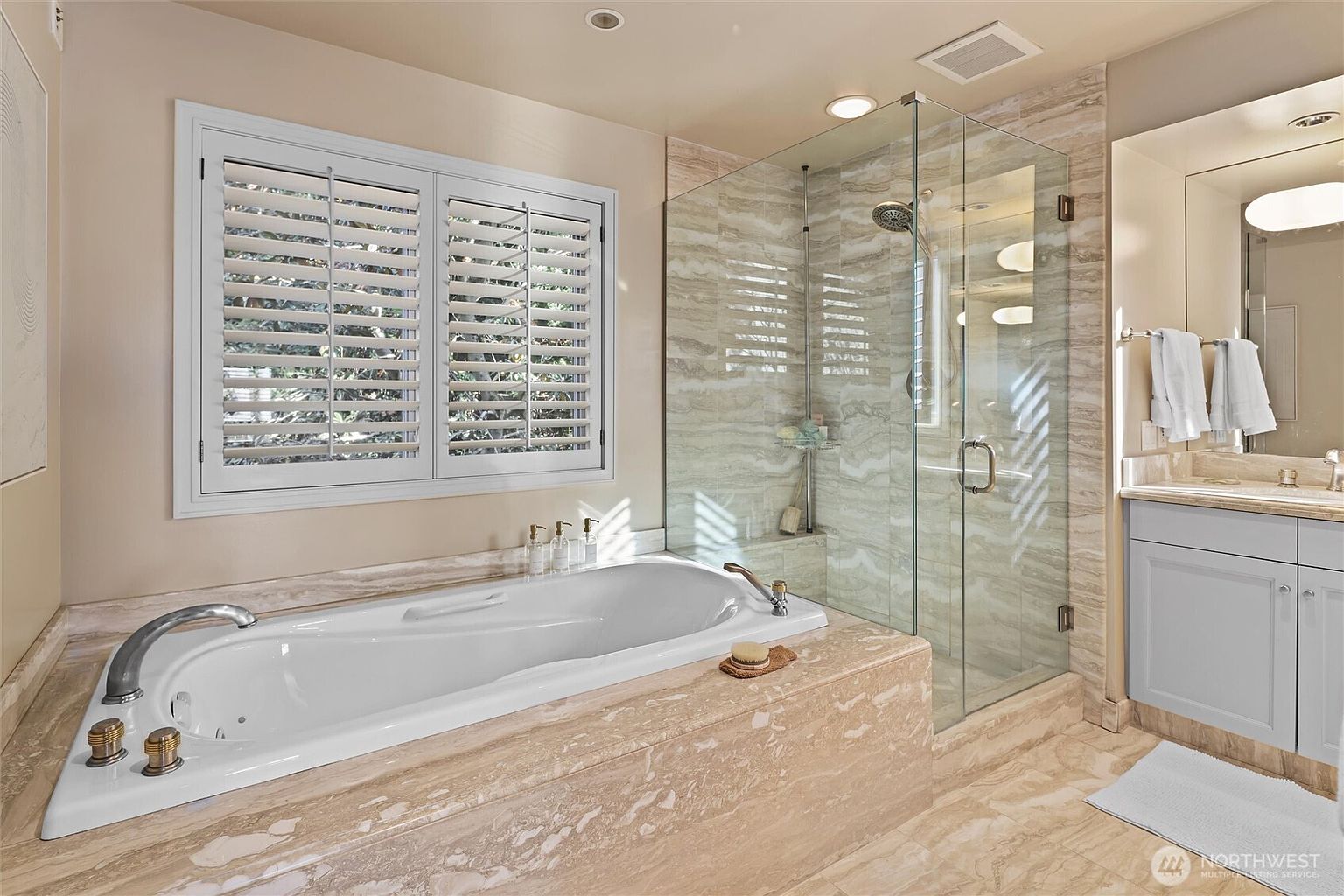 This luxurious primary bathroom features a large soaking tub set into a beige marble surround, positioned beneath a window with white plantation shutters. Adjacent to the tub is a spacious walk-in glass shower with matching marble tile work, creating a cohesive and elegant aesthetic. The vanity area includes a light-colored cabinet and a mirror, contributing to a bright, spa-like atmosphere.