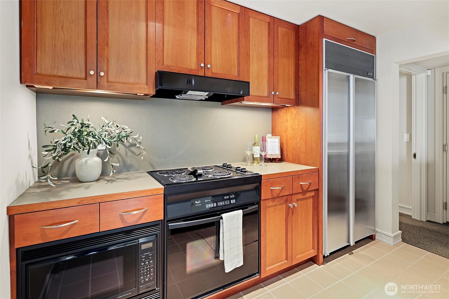This compact, functional kitchen features warm-toned wooden cabinetry paired with a sleek, matte-finish countertop and a neutral backsplash. The space is equipped with a black electric range, a built-in microwave, and a tall, stainless steel refrigerator, creating a cohesive and efficient cooking area. The perspective is a straight-on, eye-level shot that highlights the organized layout and the inviting, well-maintained aesthetic of the home.