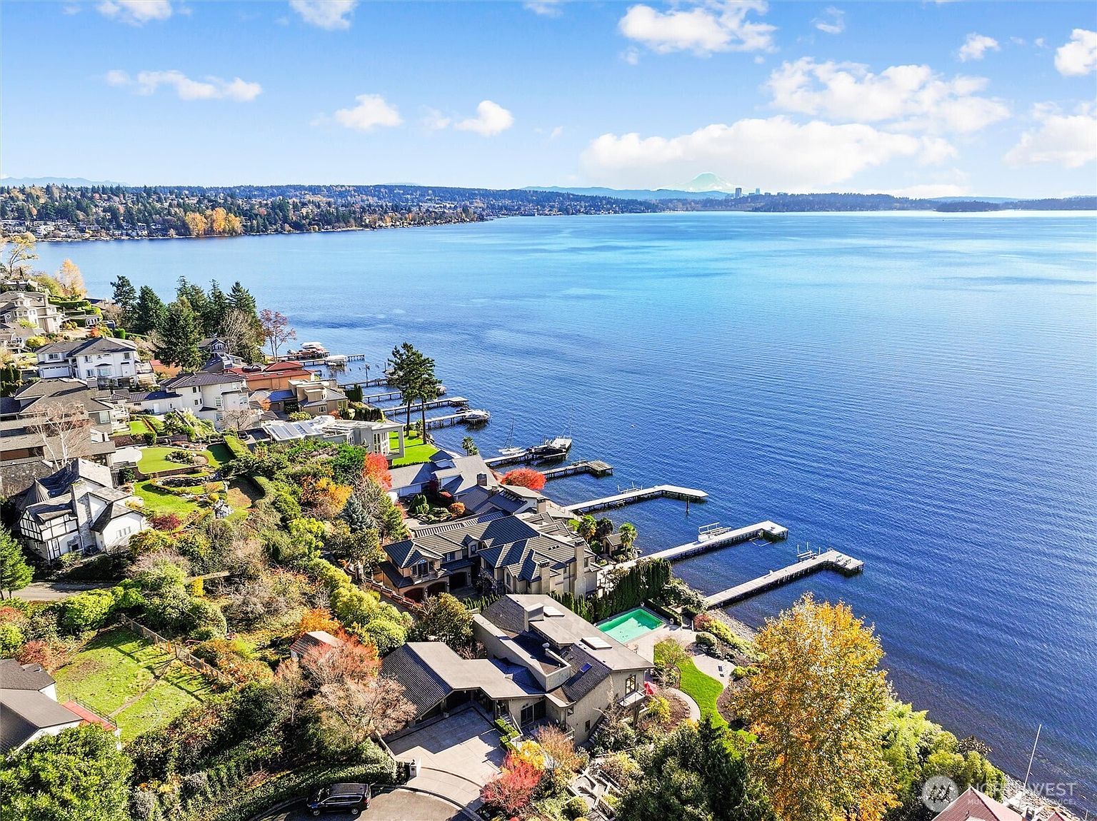 This high-angle aerial view captures a luxurious waterfront estate nestled along a serene lake, showcasing its expansive property, private dock, and swimming pool. The architecture features a multi-level design with dark roofing that blends seamlessly into the lush, tree-lined landscape. In the distance, a majestic mountain peak rises above the horizon, enhancing the picturesque and tranquil atmosphere of this prime real estate location.